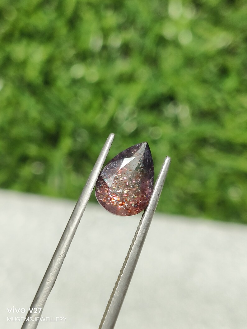 May include: A pear-shaped gemstone with a reddish-brown hue and iridescent sparkle. The gemstone is held by tweezers against a blurred green background.