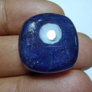May include: A deep blue, square-shaped gemstone with a rounded top. The stone has a white, circular inclusion in the center and a speckled, glittery appearance. The gemstone is held in a person's hand.