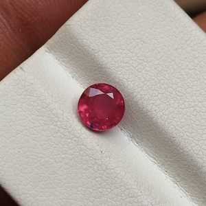 May include: A vibrant red gemstone, likely a ruby, is displayed on a white surface. The gemstone is round and has a smooth, polished finish.