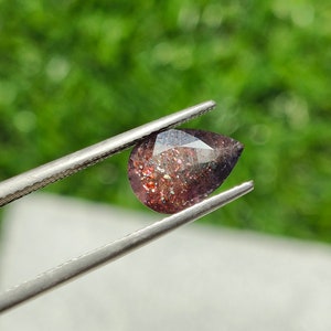 May include: A pear-shaped gemstone with a reddish-brown color and iridescent sparkle. The gemstone is held by tweezers against a green background.