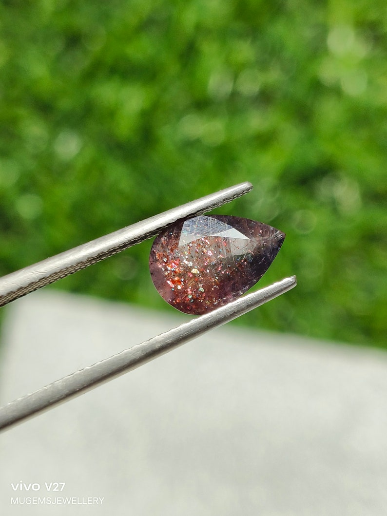 May include: A pear-shaped gemstone with a reddish-brown color and iridescent sparkle. The gemstone is held by tweezers against a green background.