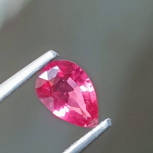 Puede incluir: Una piedra preciosa rosa en forma de pera con facetas, sostenida por unas pinzas.