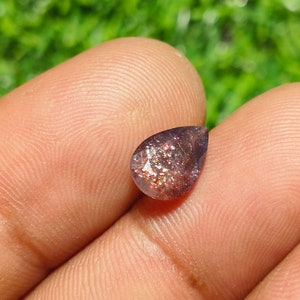 May include: A teardrop-shaped sunstone gemstone with a deep purple hue and iridescent sparkle. The stone is set against a blurred green background.