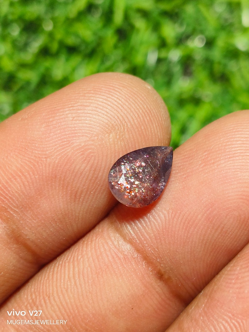 May include: A teardrop-shaped sunstone gemstone with a deep purple hue and iridescent sparkle. The stone is set against a blurred green background.