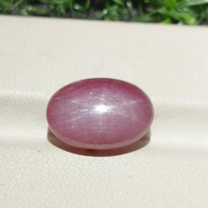 May include: A polished, oval-shaped gemstone in a deep reddish-purple hue. The stone has a smooth surface with subtle internal light reflections. The gem is set against a neutral background, highlighting its color and form. This gemstone could be used in jewelry.