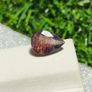 May include: A pear-shaped gemstone with a reddish-brown and purple color. The gemstone has a sparkly, iridescent finish.