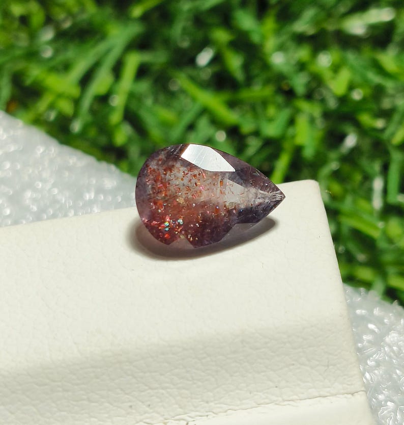 May include: A pear-shaped gemstone with a reddish-brown and purple color. The gemstone has a sparkly, iridescent finish.