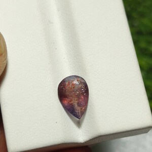May include: A teardrop-shaped gemstone with a reddish-brown and purple color. The gemstone has a sparkly, iridescent finish.