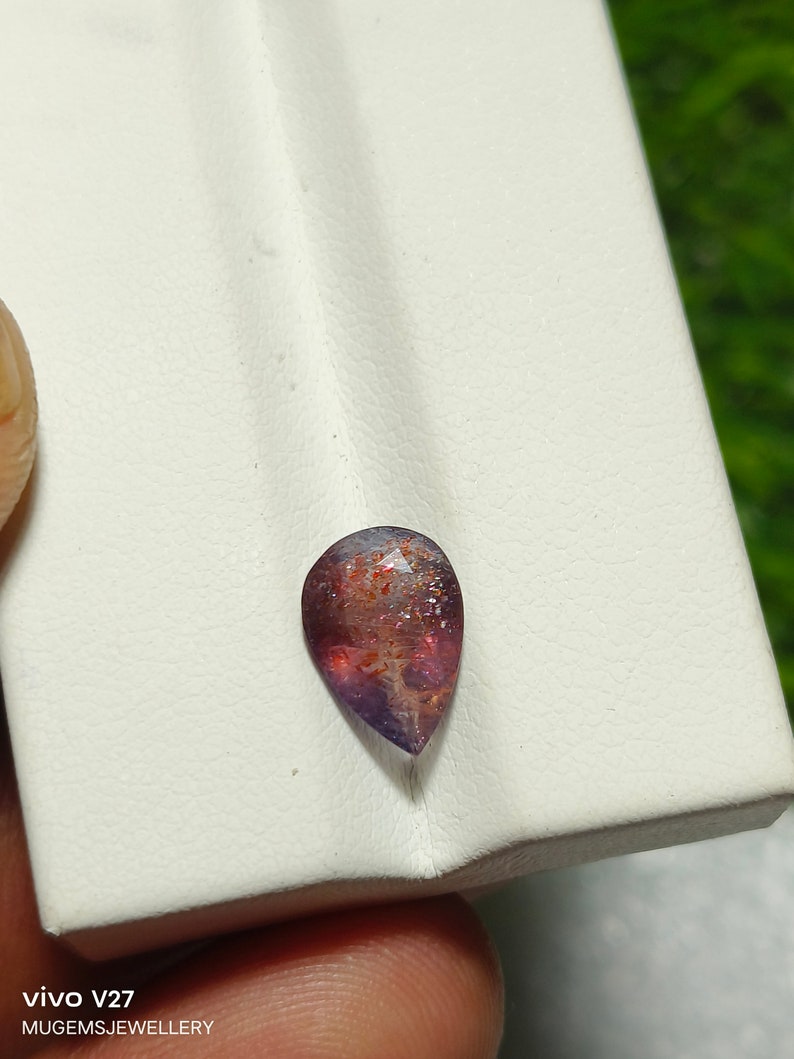 May include: A teardrop-shaped gemstone with a reddish-brown and purple color. The gemstone has a sparkly, iridescent finish.