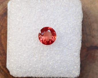May include: A round, red gemstone with a slight orange hue. The gemstone is set on a white background.