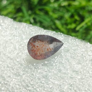 May include: A teardrop-shaped gemstone with a dark purple hue and flecks of orange and red.