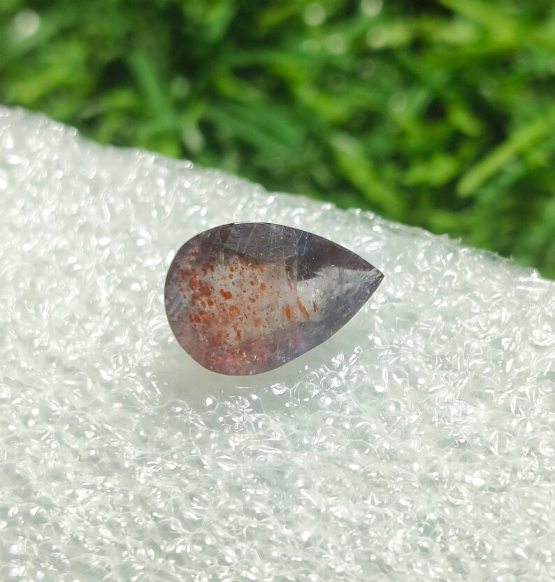 May include: A teardrop-shaped gemstone with a dark purple hue and flecks of orange and red.
