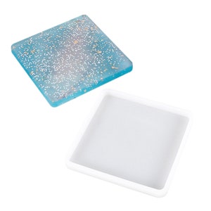 May include: Two square silicone moulds for making resin coasters. One mould is filled with blue resin with gold and white glitter. The other mould is empty.