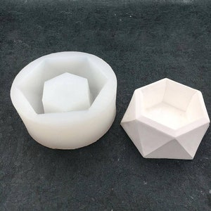 May include: A white geometric concrete planter with a silicone mold. The planter has a hexagonal shape with a flat base and a wide opening at the top. The mold is a round silicone form with a hexagonal opening in the center.