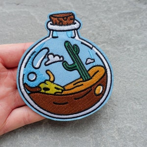 Könnte beinhalten: Eine blaue Glasflasche mit einem Korkverschluss, die eine kleine Wüstenlandschaft mit einem Kaktus, Sand und einem kleinen Tier enthält. Die Flasche ist mit braunem, grünem und gelbem Garn bestickt.