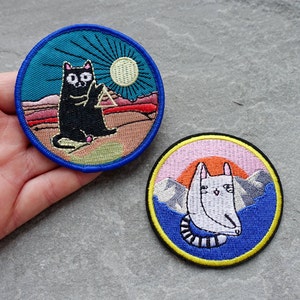 May include: Two embroidered iron-on patches featuring cats. The first patch is blue, green, and orange with a black cat holding a triangle. The second patch is blue, pink, and yellow with a white cat with a striped tail.
