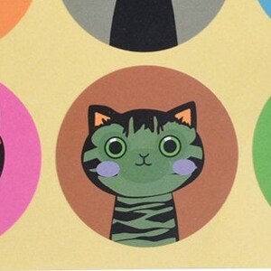 May include: A green and black striped cat with large, round eyes and pink cheeks. The cat is looking directly at the viewer. The cat is on a brown circle with a yellow background.