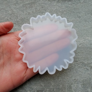 Puede incluir: Un molde de silicona transparente con forma de flor y borde ondulado. El molde se sostiene en una mano, mostrando su cualidad translúcida. Ideal para crear posavasos de resina o piezas decorativas.