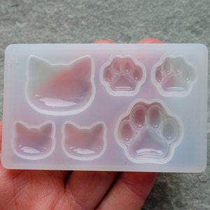 Puede incluir: Un molde de silicona transparente con caras de gato y huellas de patas. El molde tiene dos caras de gato pequeñas, una cara de gato grande y tres huellas de patas. El molde se sostiene en una mano.