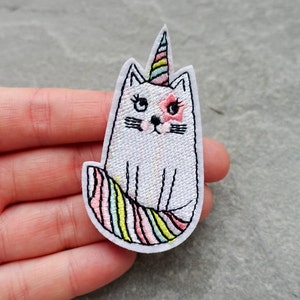 May include: White embroidered cat with a pink and yellow unicorn horn, sitting on a rainbow striped tail.