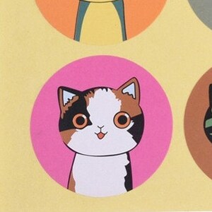 May include: A pink circle sticker with a cartoon illustration of a calico cat with brown, white, and black fur. The cat has big, round, green eyes and a pink tongue sticking out.