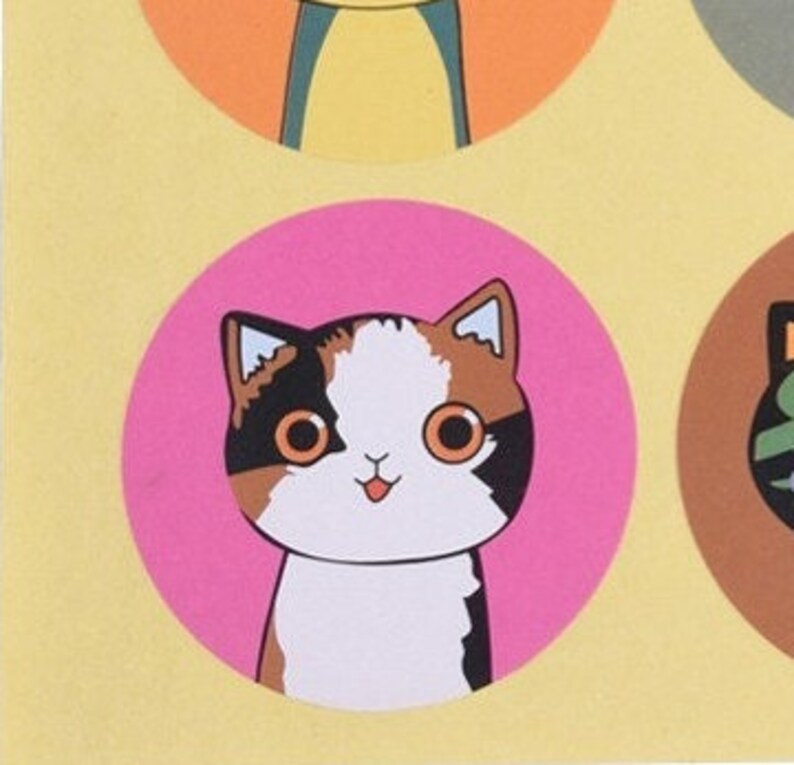 May include: A pink circle sticker with a cartoon illustration of a calico cat with brown, white, and black fur. The cat has big, round, green eyes and a pink tongue sticking out.