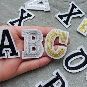May include: Close-up of three iron-on patches in the shape of the letters A, B, and C. The letter A is black with a white outline, the letter B is silver with a white outline, and the letter C is gold with a white outline.