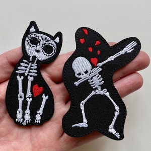 Puede incluir: Dos parches bordados para planchar que representan figuras de esqueletos. Un parche muestra un gato esqueleto con un corazón rojo. El otro parche muestra un esqueleto haciendo el dab con corazones rojos alrededor.