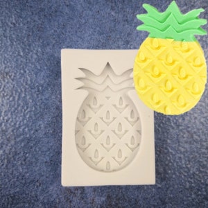 May include: A white silicone pineapple mold next to a yellow and green pineapple. The mold is in the shape of a pineapple with detailed grooves. The pineapple is yellow with a green crown. The background is a textured blue surface.