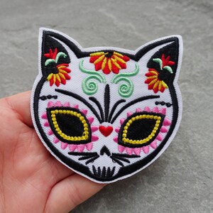 May include: White embroidered iron-on patch of a cat with a Day of the Dead sugar skull design. The cat's eyes are black with yellow accents, and the skull is decorated with pink, red, yellow, and green flowers.