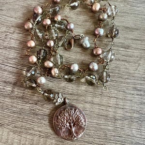 May include: A handcrafted necklace featuring a pendant with a tree design. The necklace combines pink pearls, clear faceted beads, and smoky quartz beads, strung on a copper-colored wire. The pendant is a circular, silver-toned metal piece.