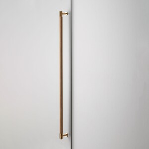 May include: A modern door pull with a wooden handle and brass-colored hardware. The long, vertical handle is made of light brown wood, and the mounting hardware is a bright gold color. The door pull is mounted on a white surface.