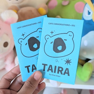 May include: Two light blue booklets with a bear illustration and the text "CAFE CONVERSATIONS .001" and "TAIRA by elisa milan". The booklets have a star and swirl design. The background includes plush toys.