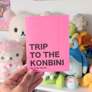 May include: A pink booklet with the title "TRIP TO THE KONBINI" in black text. The text "by ELISA MILAN" is printed below the title.