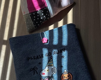 Pode incluir: Uma bolsa de ganga azul-escura com fecho de botão de flor rosa e um gráfico bordado de uma personagem com chapéu de festa a ler um livro ao lado de uma abóbora, com o texto PLEASE GO AWAY. Por cima, encontra-se uma bolsa de tecido patchwork.