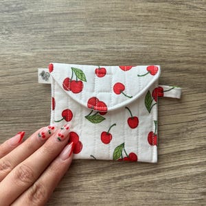 May include: A small white quilted pouch with a cherry print. The pouch has a flap closure and a small loop handle.