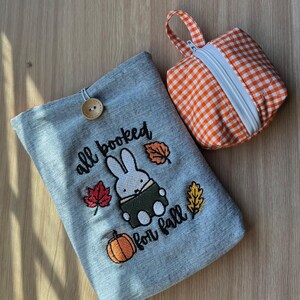 Pode incluir: Uma capa de livro de ganga com um coelho bordado, uma ab&oacute;bora e folhas de outono, com o texto "all booked for fall". Uma pequena bolsa com z&iacute;per xadrez laranja e branco com uma al&ccedil;a tamb&eacute;m est&aacute; presente.
