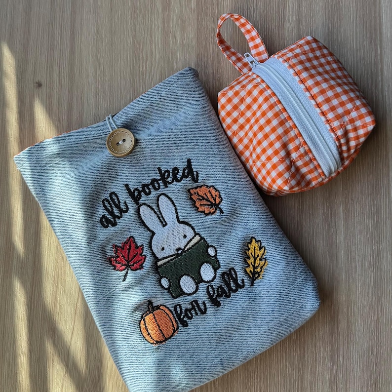 Pode incluir: Uma capa de livro de ganga com um coelho bordado, uma ab&oacute;bora e folhas de outono, com o texto "all booked for fall". Uma pequena bolsa com z&iacute;per xadrez laranja e branco com uma al&ccedil;a tamb&eacute;m est&aacute; presente.