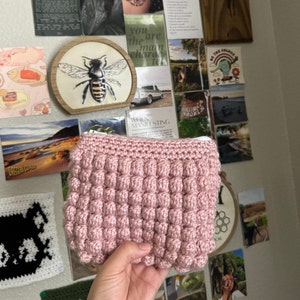 May include: A pink crocheted pouch with a zipper closure. The pouch is made with a textured stitch that creates a bubble-like effect.