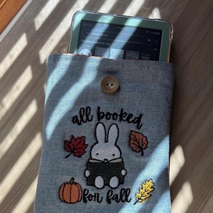 May include: A denim Kindle sleeve with a button closure. The sleeve features an embroidered design with the text "all booked for fall", a cartoon rabbit reading a book, and fall-themed leaves and a pumpkin.