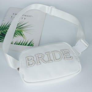 May include: White fanny pack with the word "BRIDE" spelled out in faux pearls. The bag has a white adjustable strap with a plastic buckle. A book with the text "THE INGU-TY" is in the background.