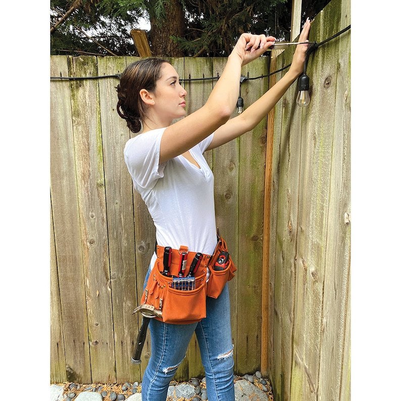 May include: A person wearing a white t-shirt and blue jeans is installing outdoor string lights. They are using a screwdriver to attach the lights to a wooden fence. The person is wearing a brown leather tool belt with various tools and pouches.