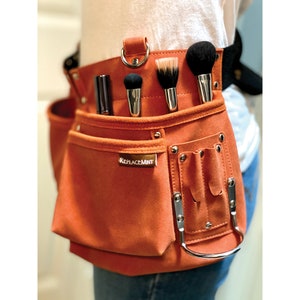 May include: Orange suede makeup artist tool belt with multiple pockets and a ReplaceMint logo. The belt has a silver ring and is worn around the waist.