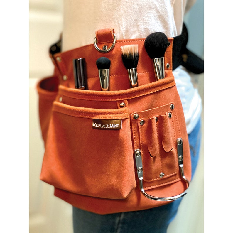 May include: Orange suede makeup artist tool belt with multiple pockets and a ReplaceMint logo. The belt has a silver ring and is worn around the waist.