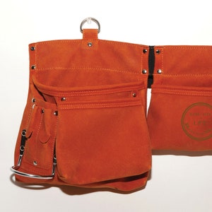 May include: A brown leather tool belt with multiple pockets and a metal ring for hanging. The belt has a black strap and metal rivets.
