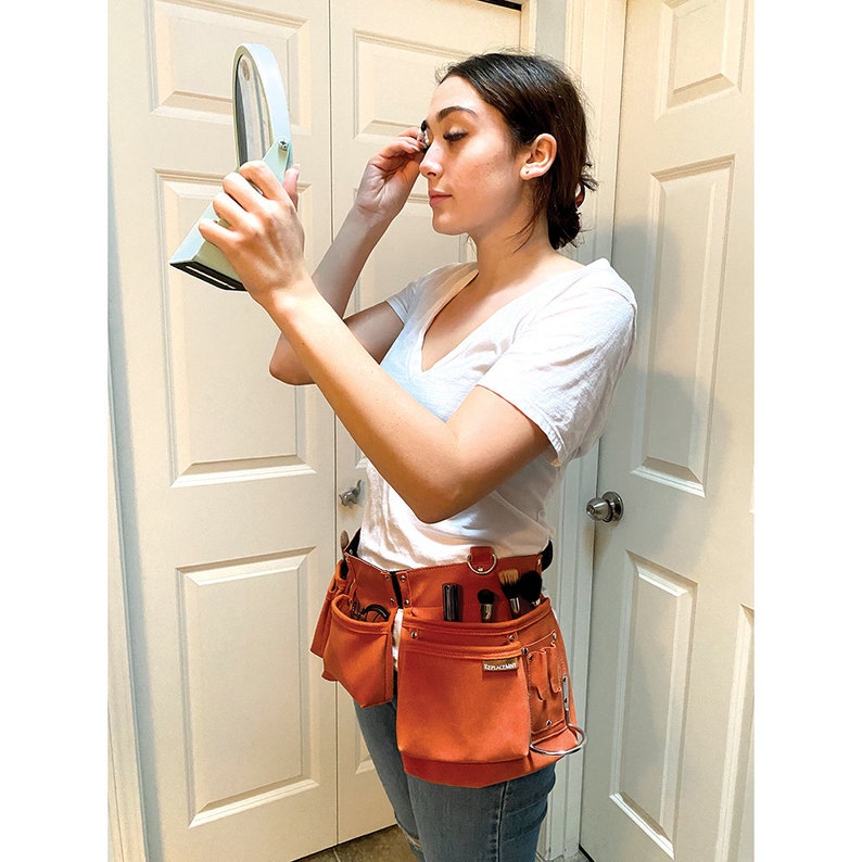 May include: A woman wearing a white t-shirt and blue jeans is using a handheld mirror to apply makeup. She is wearing a brown leather tool belt with multiple pockets and tools.