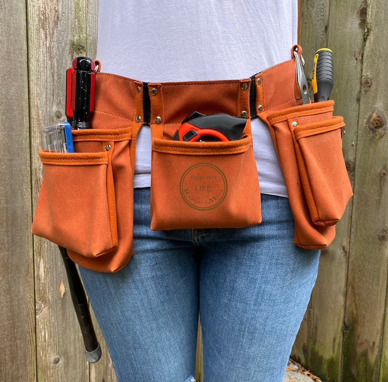 May include: A brown canvas tool belt with multiple pockets, including a large pocket with the text "Enjoy Life" in a circle. The belt is worn over blue jeans.