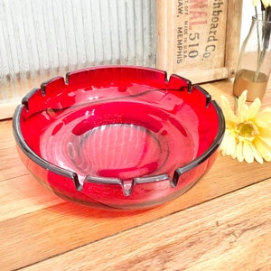 May include: A red glass ashtray with a wavy design. The ashtray has four indentations around the rim.