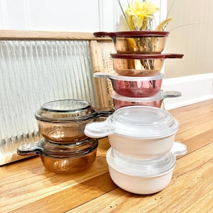 May include: A stack of vintage glass and ceramic bowls with lids. The bowls are in various colors, including brown, white, and pink. The bowls are stacked on a wooden surface.