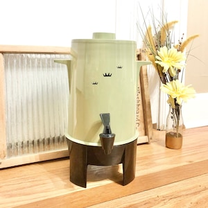 May include: A vintage green and black coffee pot with a black spout and a black base. The pot has a design of three black crowns on the front.